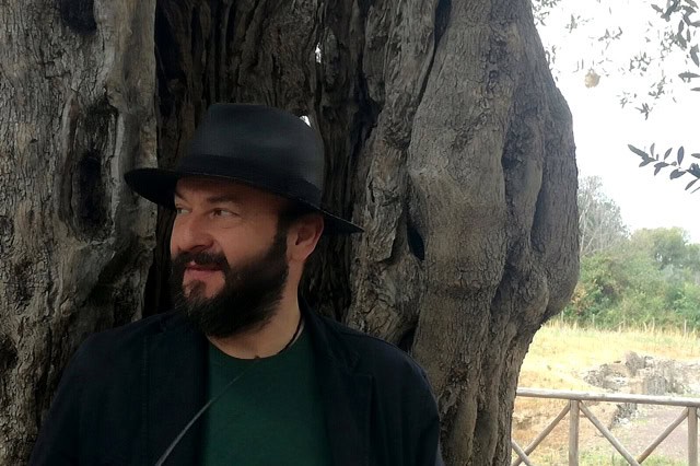 Uomo con cappello nero e barba sotto al albero in campagna.